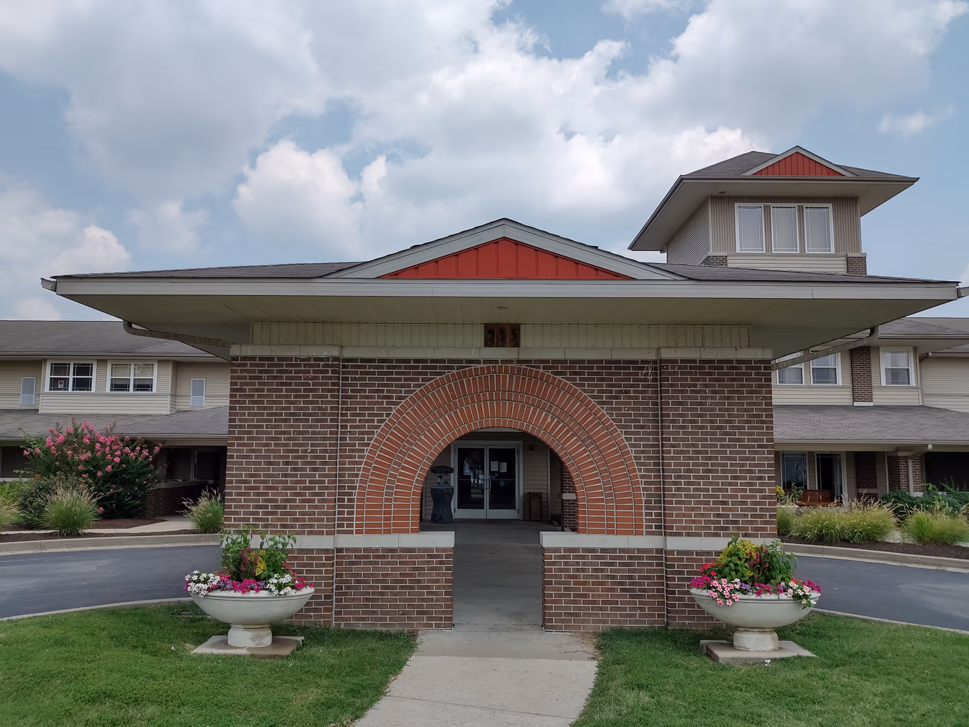 Front exterior view of a senior living facility with a brick archway entrance, two large flower planters on either side, and a multi-story building with windows and a gray roof under a partly cloudy sky.