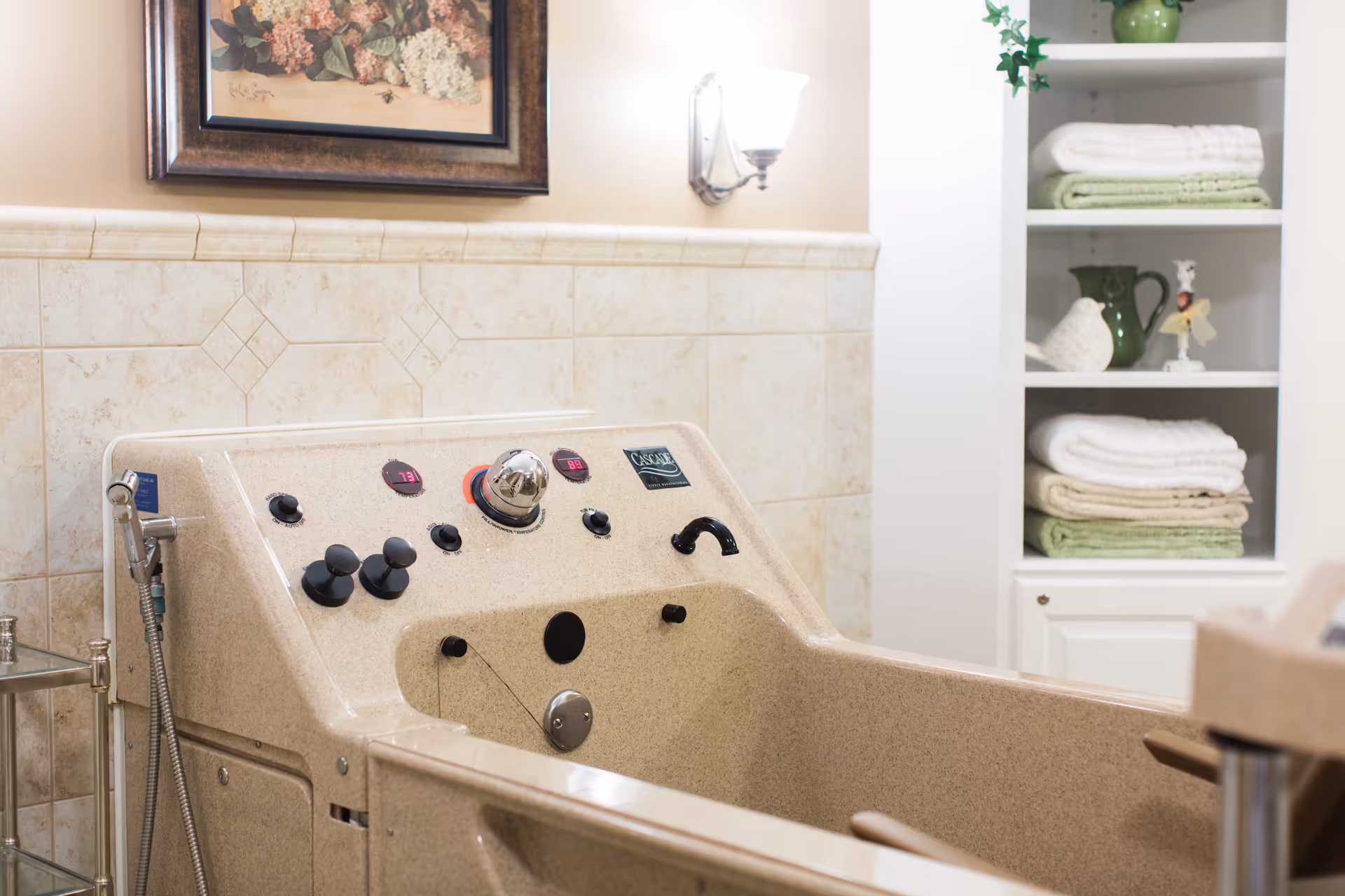 A beige therapeutic bath tub with control knobs and temperature displays in a tiled bathroom. A wall-mounted light fixture and a framed floral painting are visible above the tub. To the right, there is a white shelving unit with neatly folded towels and decorative items.