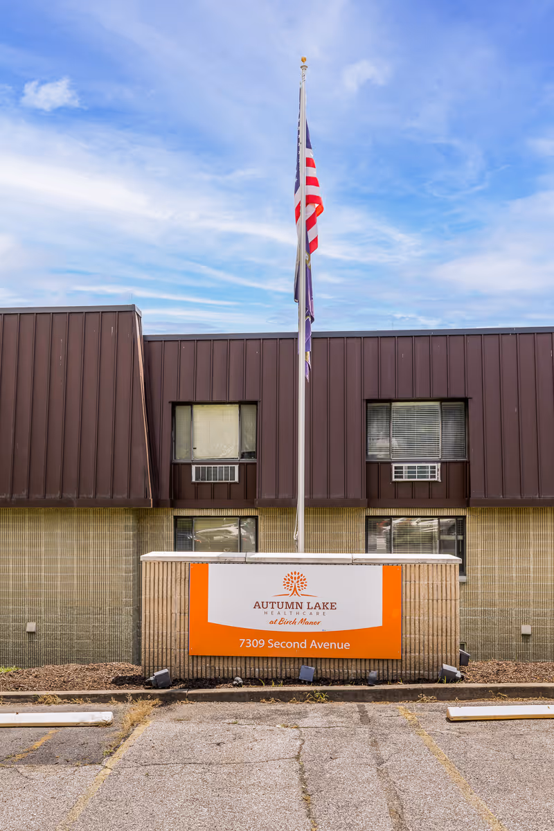 Exterior front view of Autumn Lake Healthcare at Birch Manor building with a sign displaying the facility name and address 7309 Second Avenue. An American flag and another flag are flying on a flagpole above the sign. The building has brown paneling and windows with air conditioning units.