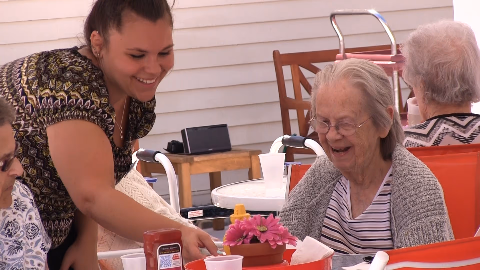A smiling young woman interacts with an elderly woman seated in an orange chair at a table outdoors. The elderly woman is laughing and wearing glasses, a striped shirt, and a gray cardigan. The table has a small potted plant with pink flowers, a ketchup bottle, and plastic cups. Other elderly individuals are seated nearby, and the setting appears to be a patio area with white siding in the background.