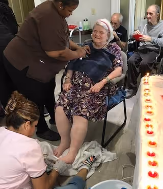An elderly woman seated in a chair receiving a foot bath from a caregiver kneeling in front of her, while another caregiver stands beside her. In the background, two elderly men are seated, one holding a red cup. A table with lit candles is visible on the right side of the image.