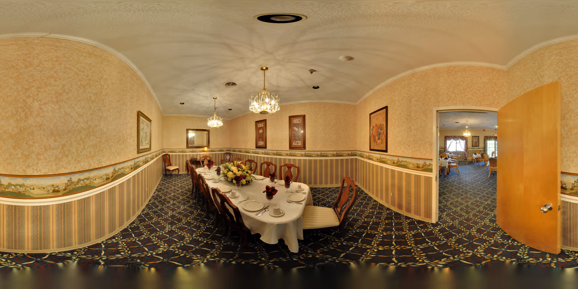 A formal dining room with a long table set for a meal, including plates, cups, and napkins. The room has patterned wallpaper with a border, carpeted floor with a floral design, and two chandeliers hanging from the ceiling. There are framed pictures on the walls and an open door leading to another dining area with more tables and chairs.