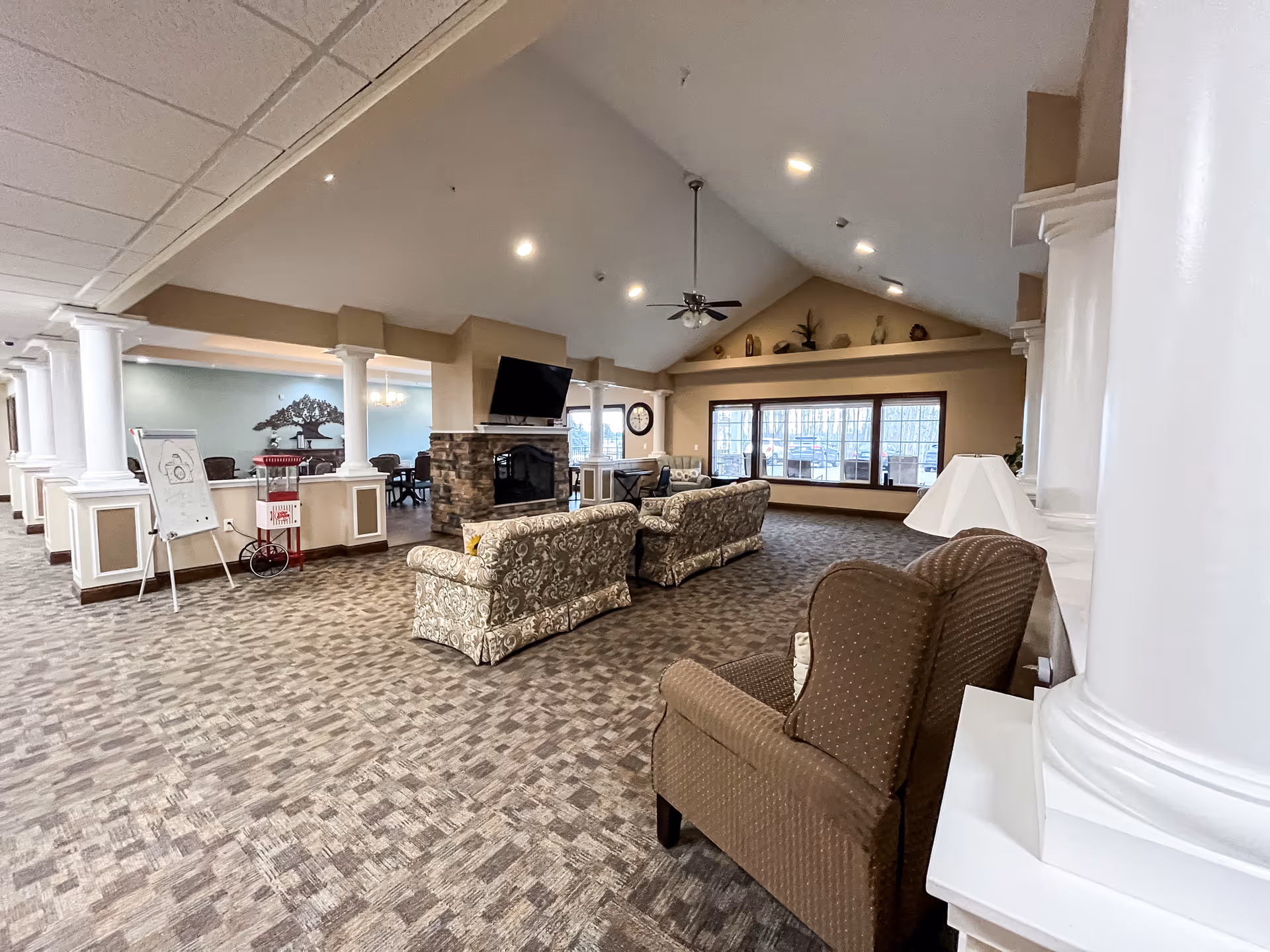 Spacious assisted living facility common area with patterned carpet, multiple upholstered sofas and armchairs arranged around a stone fireplace with a mounted TV. Large windows let in natural light, and decorative columns separate this space from an adjacent dining area with tables and chairs. The ceiling is vaulted with recessed lighting and a ceiling fan.