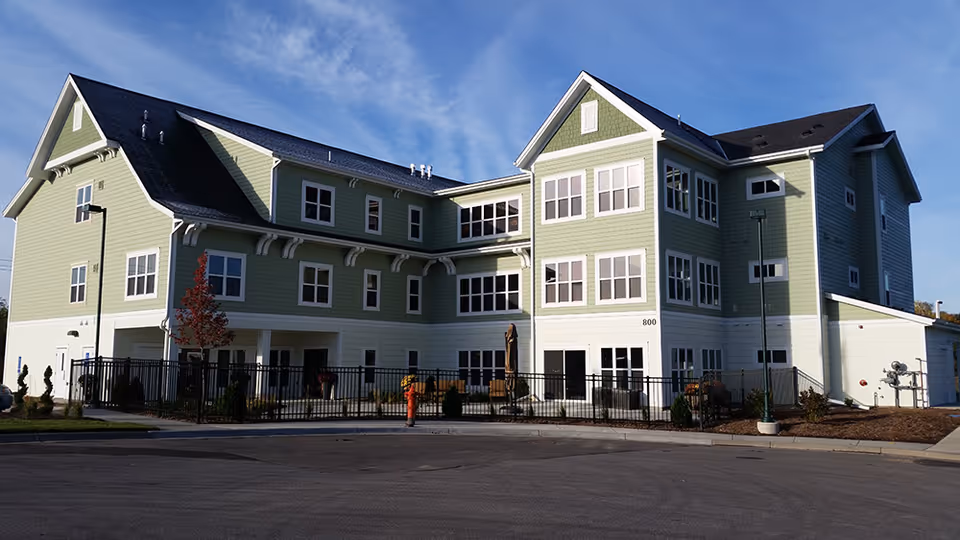 Exterior view of a three-story green and white senior living facility building with multiple windows, a black fence surrounding a small garden area, and a clear blue sky in the background.