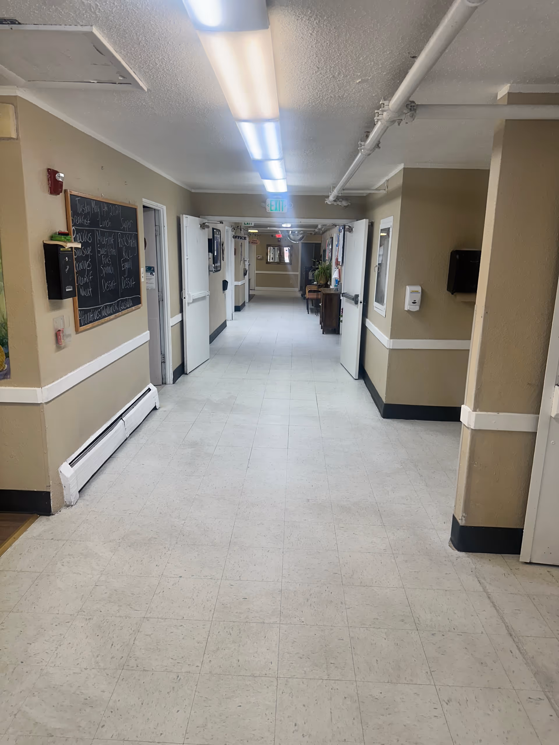 A long, clean hallway in an assisted living facility with beige walls, white tiled floor, and fluorescent ceiling lights. There are open double doors on the right and left sides, a bulletin board on the left wall, and an exit sign at the far end of the corridor.