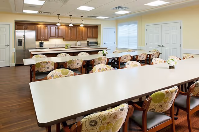 A bright dining room with long white tables and wooden chairs featuring patterned cushions. The room has a kitchen area in the background with wooden cabinets, a refrigerator, a microwave, and a coffee maker. The walls are painted light yellow, and there are windows with blinds letting in natural light.