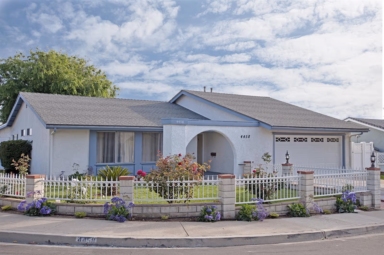 Single-story light-gray suburban house with a fenced front yard, garage, and flowering plants under a partly cloudy sky.