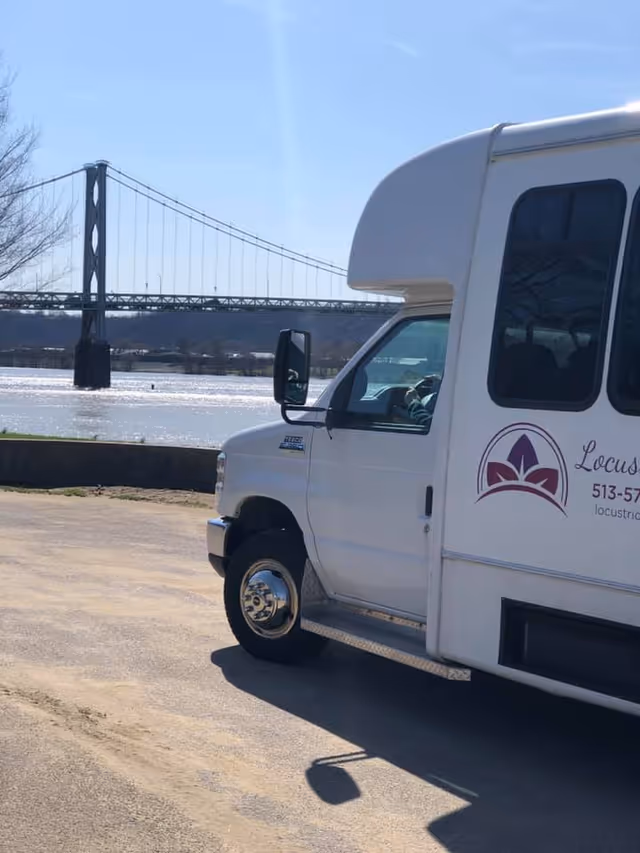 Partial view of a white shuttle bus with Locust Ridge Nursing and Rehabilitation logo and contact information on the side, parked near a body of water with a suspension bridge in the background under a clear blue sky.
