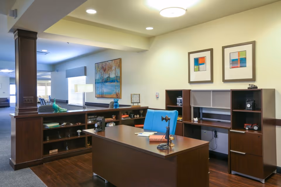 A well-lit office area in Orchard Grove Senior Living featuring a wooden desk with a blue chair, a desk lamp, books, and decorative items. Behind the desk is a wooden shelving unit with various small decorative objects and two framed abstract paintings on the wall. The space has a warm and inviting atmosphere with soft yellow walls and wood flooring.