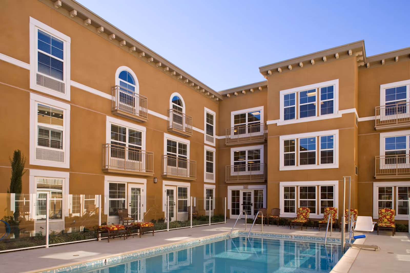 Outdoor swimming pool area at Belmont Village Senior Living Cardiff by the Sea, surrounded by a three-story building with multiple windows and small balconies. Poolside chairs with colorful cushions are arranged along the pool deck, and a clear safety fence encloses the pool.