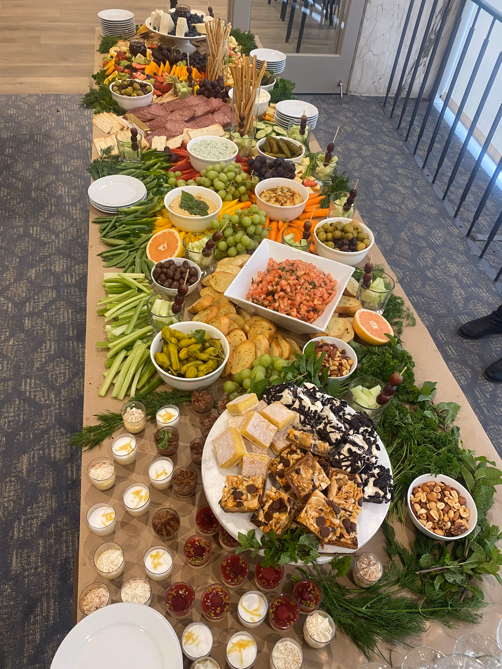 A long table set with a variety of appetizers and snacks including fresh vegetables, grapes, olives, crackers, dips, sliced meats, nuts, and assorted desserts. The table is decorated with greenery and plates are stacked at one end. The setting appears to be indoors with carpeted flooring and a railing visible on one side.