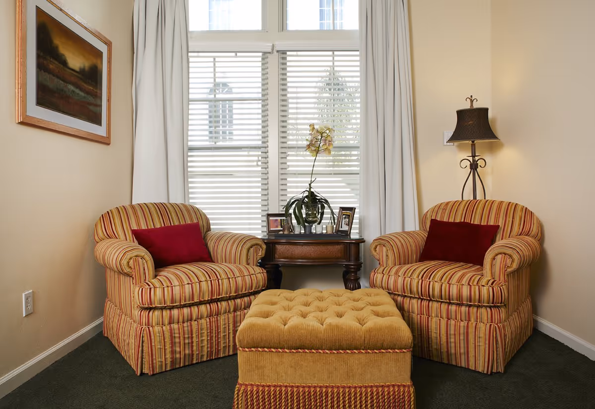 Two striped armchairs with red pillows face a tufted ottoman in a cozy sitting area by a window with a side table and lamp.