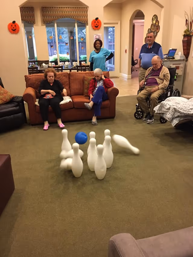 A group of elderly people and a caregiver in a living room area playing indoor bowling. Two elderly women are seated on a brown couch, one elderly man is in a wheelchair, and another man stands nearby. The bowling pins are in motion with a blue ball hitting them. The room is decorated with Halloween pumpkin decorations on the walls.