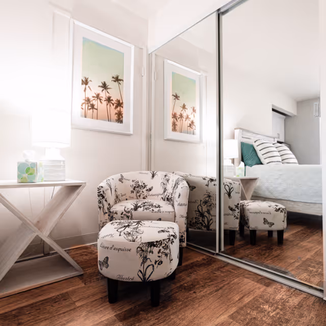 A cozy corner of a bedroom featuring a patterned armchair with a matching ottoman on wooden flooring. Next to the chair is a white side table with a lamp and a box of tissues. A framed picture of palm trees hangs on the wall above the chair. A large mirrored sliding closet door reflects the chair and part of the bed with striped pillows in the background.