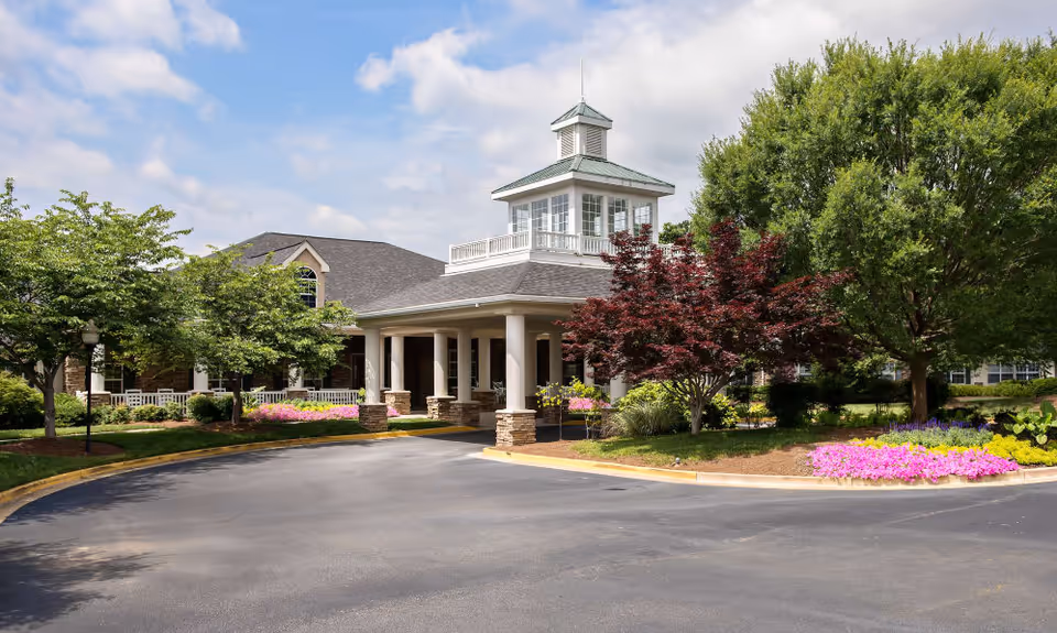 Exterior view of Azalea Estates of Fayetteville showing the main entrance with a covered driveway, surrounded by well-maintained landscaping including trees, bushes, and colorful flower beds under a partly cloudy sky.