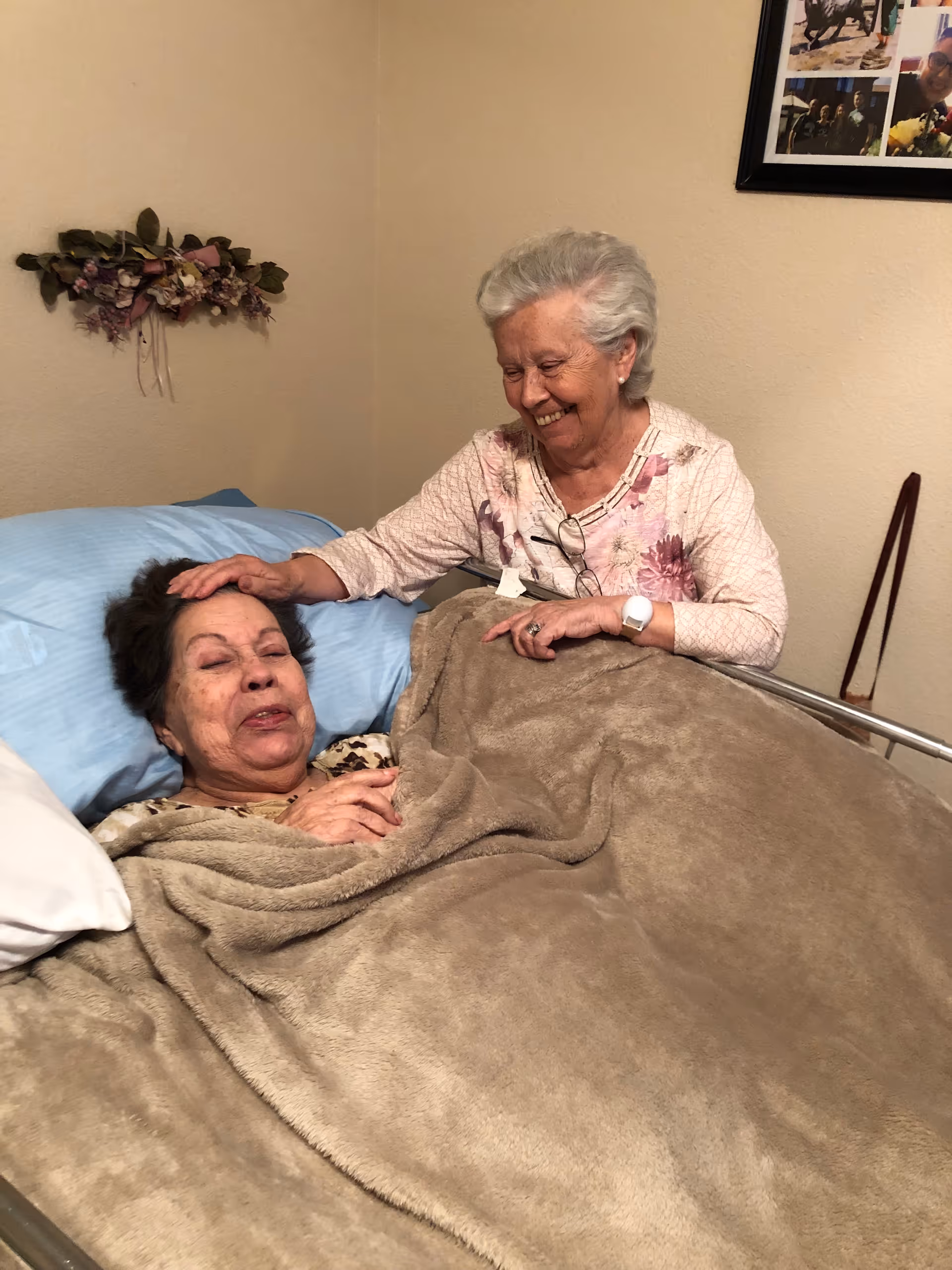 An elderly woman lying in a bed covered with a beige blanket, smiling with her eyes closed, while another elderly woman sits beside her, gently touching her head and smiling warmly. The room has beige walls with a floral decoration and a framed photo collage on the wall.