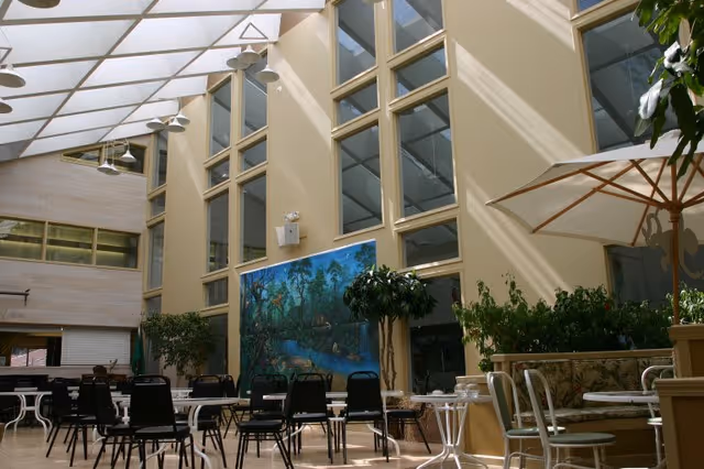A bright indoor common area with high ceilings and large windows letting in natural light. The space features several tables and chairs arranged for seating, potted plants, and a large mural on the wall depicting a nature scene. There is also a large umbrella providing shade over one seating area.