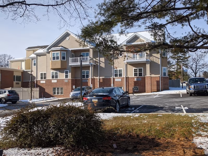 Exterior view of a multi-story residential building with beige siding and red brick accents. Several cars are parked in the parking lot in front of the building. There are leafless trees and patches of snow on the ground, indicating a winter setting.