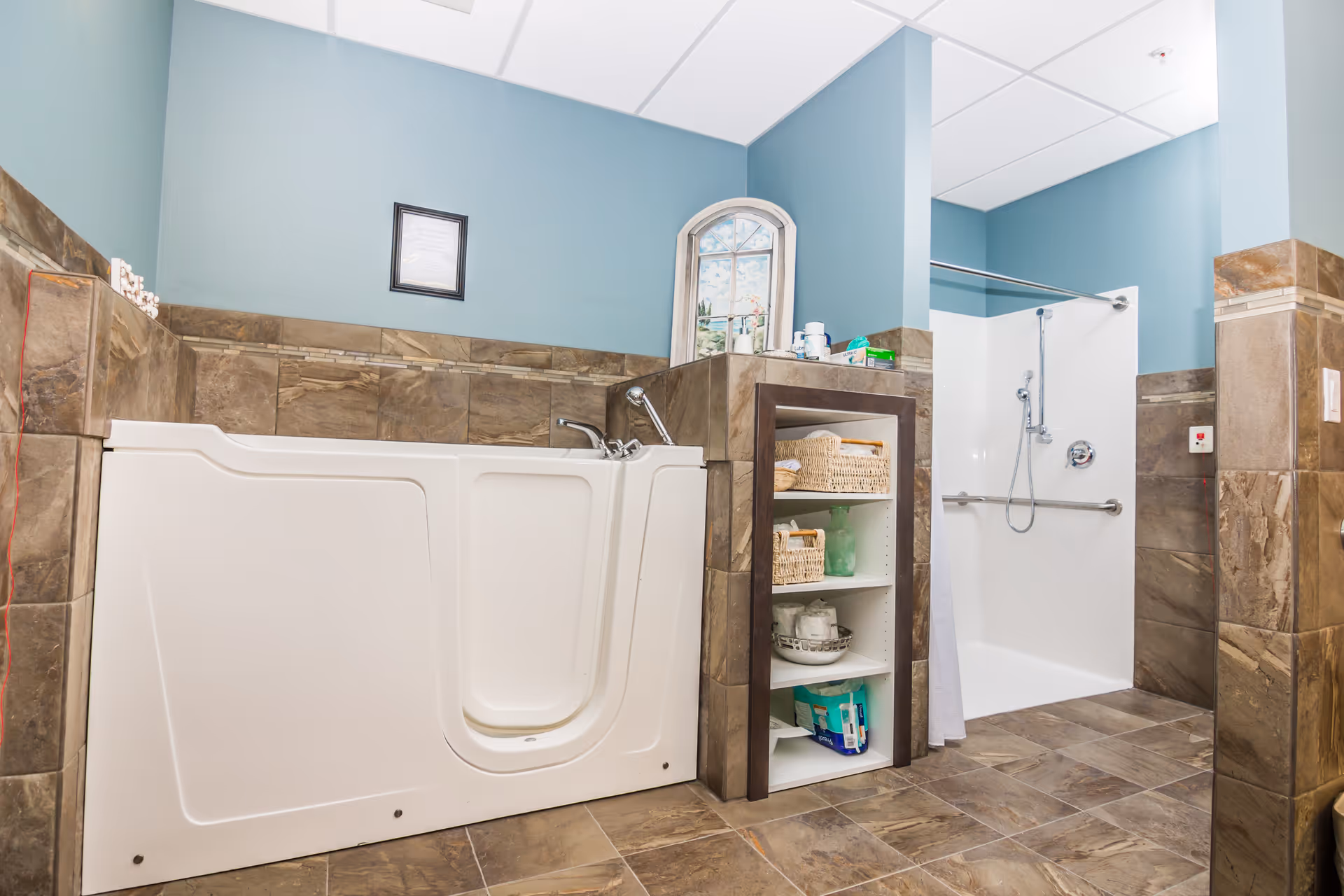 A bathroom with a walk-in bathtub and a walk-in shower. The walls are painted blue with brown tile halfway up. There is a small shelving unit between the tub and shower holding baskets and toiletries. A framed picture and a decorative window are on the wall above the tub.