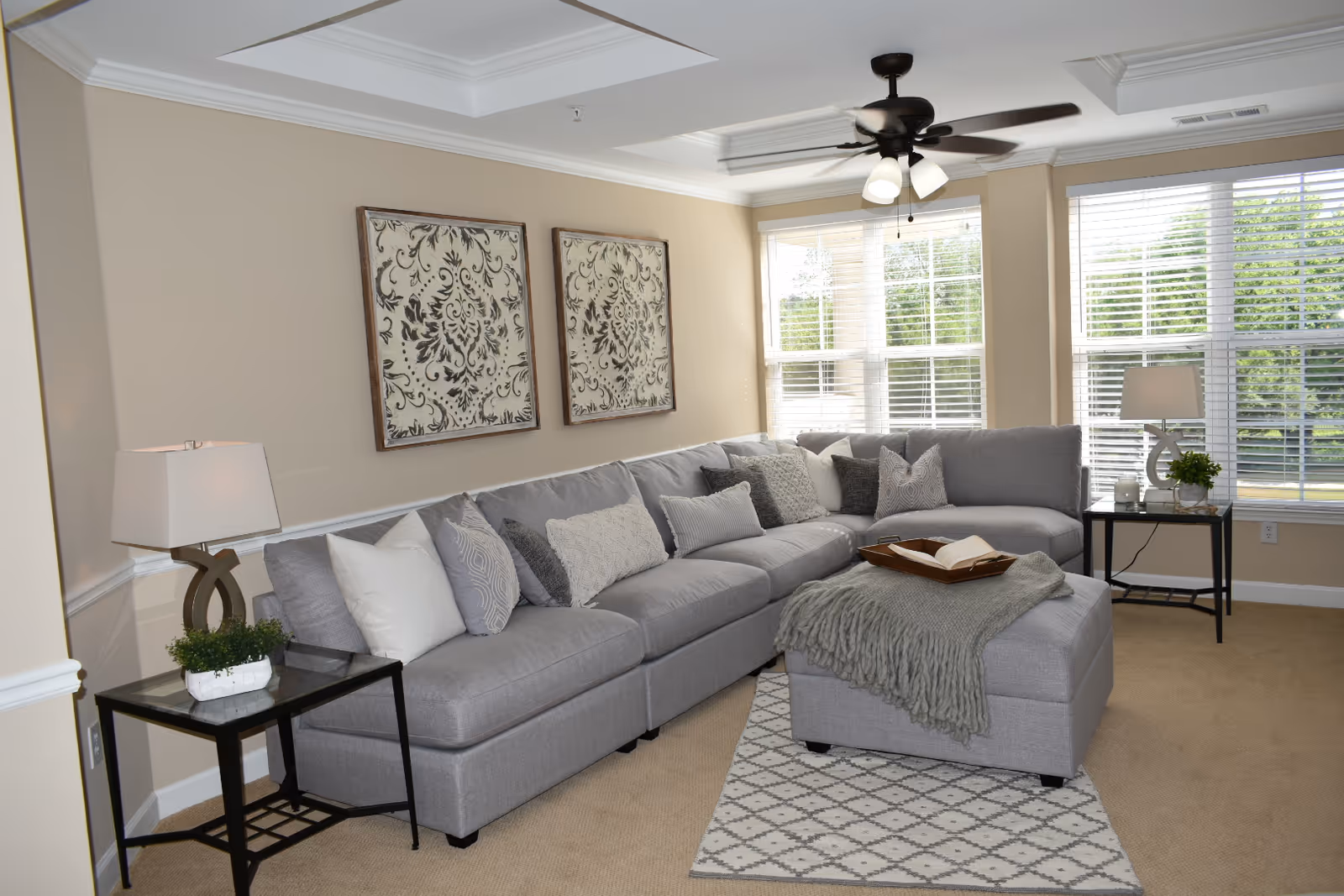 Bright living room with a gray sectional sofa and ottoman, decorative pillows, side tables with lamps, large windows with white blinds, and a ceiling fan.