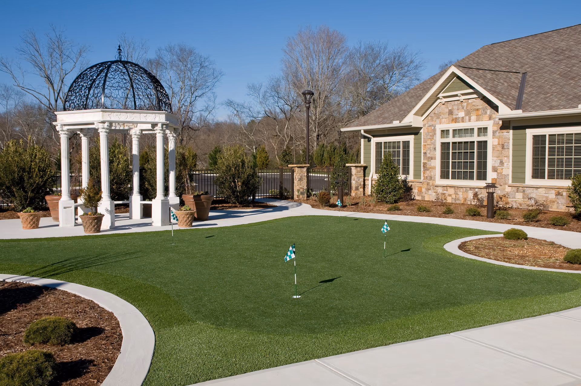 Outdoor area of a senior living facility featuring a small putting green with three golf flags, a white gazebo with ornate black metal dome roof, potted plants, and a building with stone and green siding in the background under a clear blue sky.