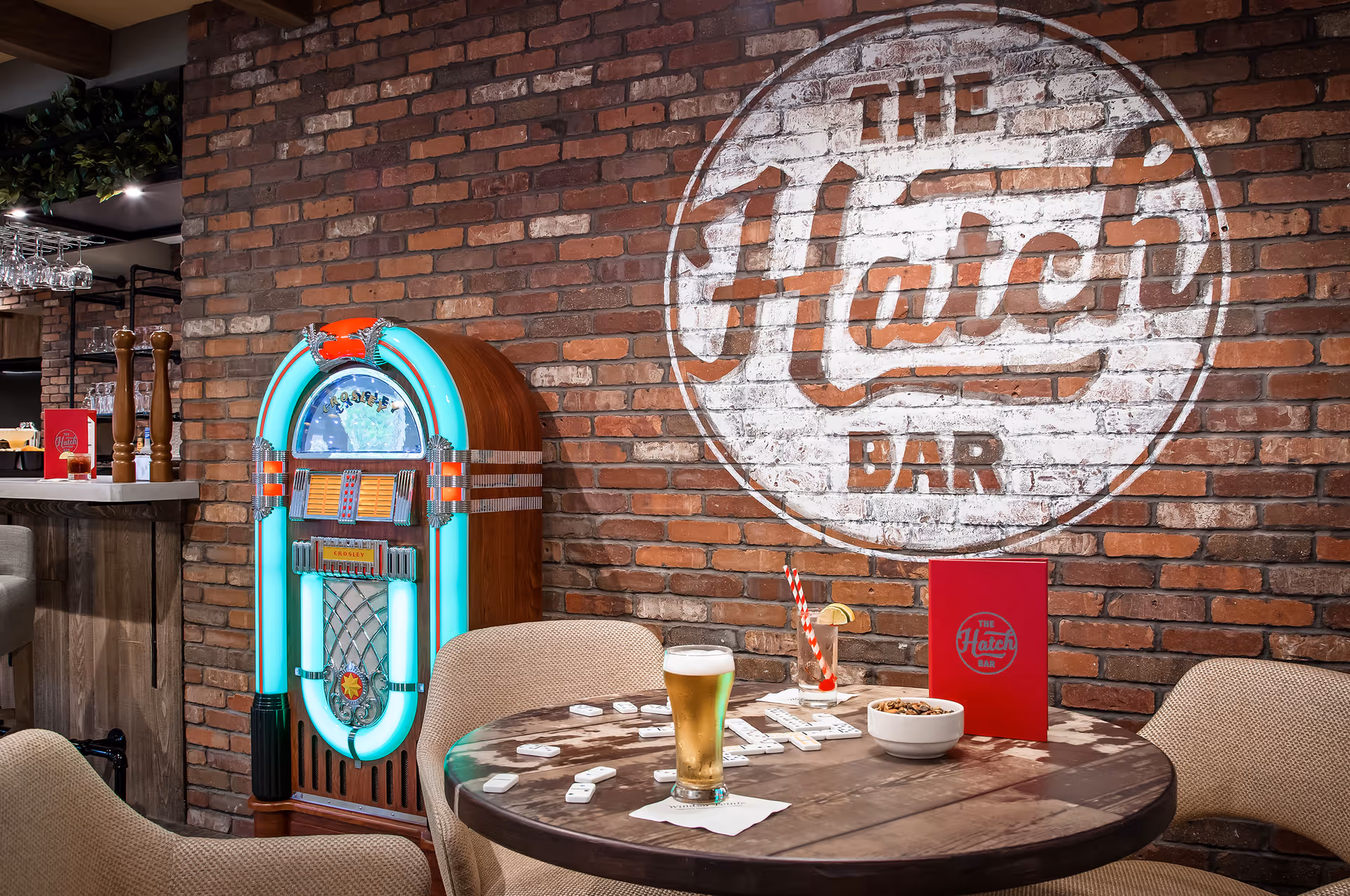 Cozy brick-walled bar area with a glowing jukebox, a round table holding a beer, snacks and dominoes, and a painted 'The Hatch Bar' logo on the wall.
