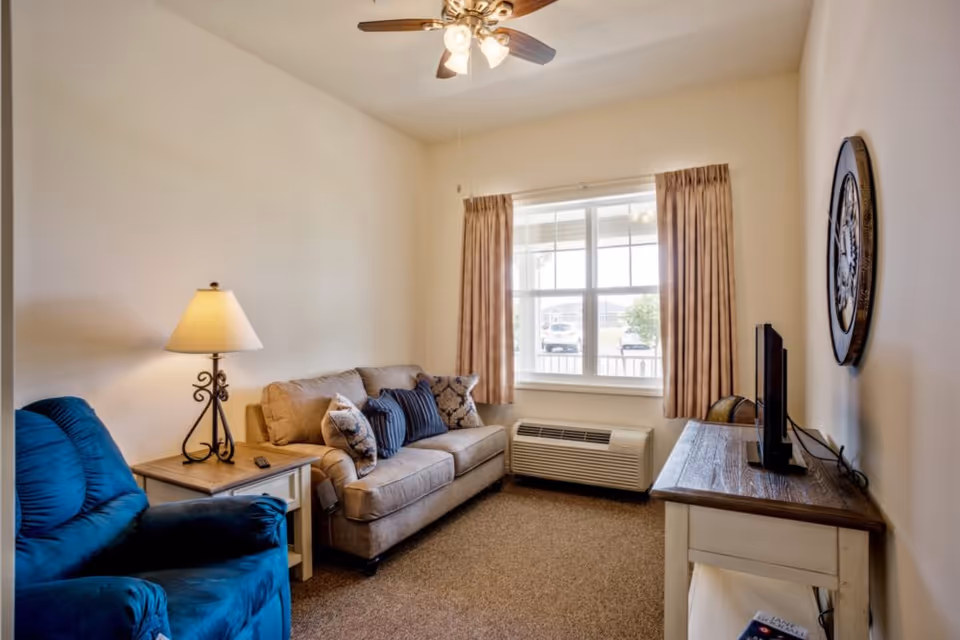 Small living room with a sofa, blue recliner, side table with lamp, TV on a console, window with curtains, and a ceiling fan.
