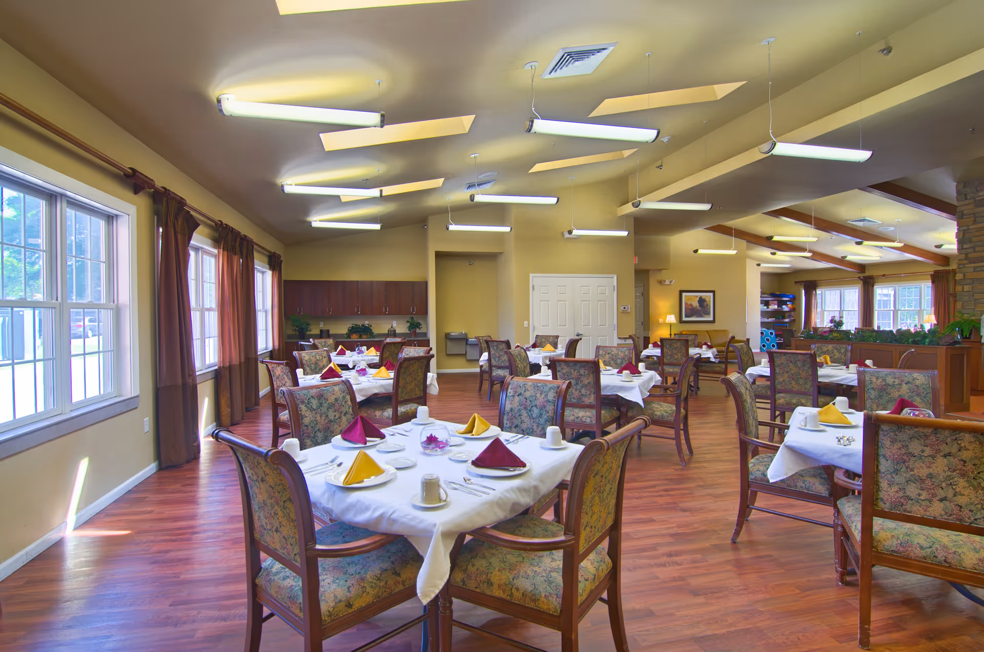 A spacious dining room in an assisted living facility with multiple tables covered in white tablecloths, each set with cups, plates, and folded napkins in yellow and burgundy colors. The room has large windows with brown curtains, wooden flooring, and ceiling lights. Floral upholstered chairs surround the tables, and there are plants and cabinets along the walls.