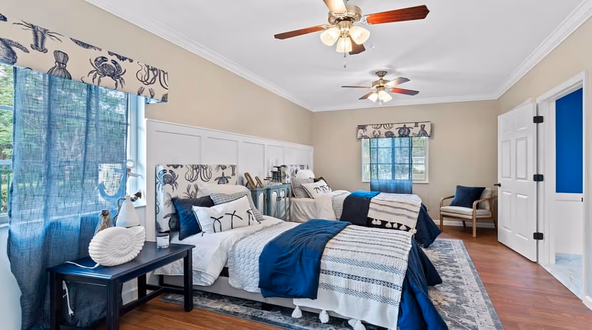 A bright bedroom with two twin beds featuring blue and white bedding and nautical-themed headboards. The room has light beige walls, two windows with blue curtains and valances decorated with sea creatures, a wooden floor, a patterned rug, a small table with decorative items, a chair with a dark blue cushion, and two ceiling fans with lights.