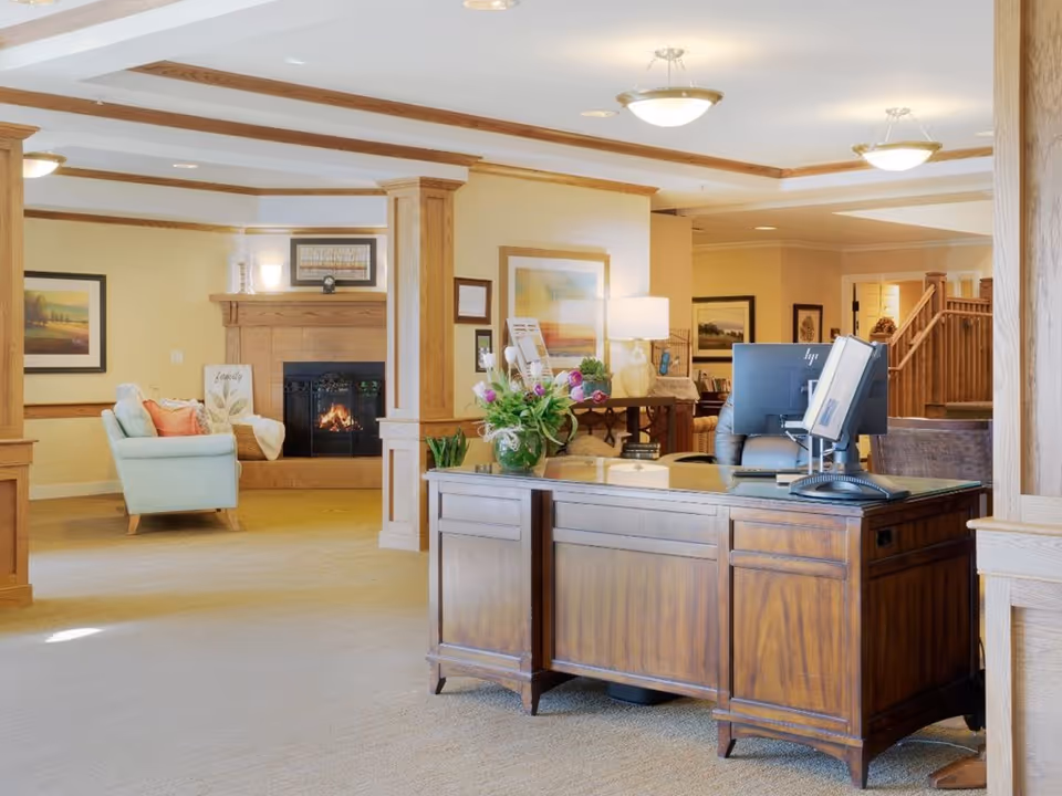 A warm and inviting interior space featuring a wooden reception desk with a computer and a vase of flowers. In the background, there is a cozy seating area with a light blue armchair, a fireplace with a wooden mantel, framed artwork on the walls, and soft lighting from ceiling fixtures and a table lamp.