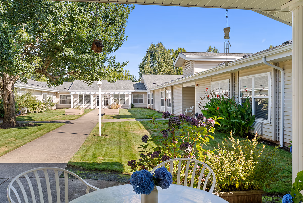 A covered patio table with chairs and flowers overlooking a landscaped courtyard surrounded by single-story senior living buildings.
