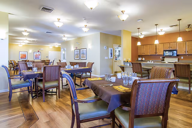 A spacious dining room in a senior living facility with multiple wooden tables and chairs arranged neatly. Tables are set with glasses, napkins, and utensils. The room has warm lighting with ceiling fixtures and pendant lights over a kitchen counter area in the background. The walls are decorated with framed artwork, and the floor is wooden.