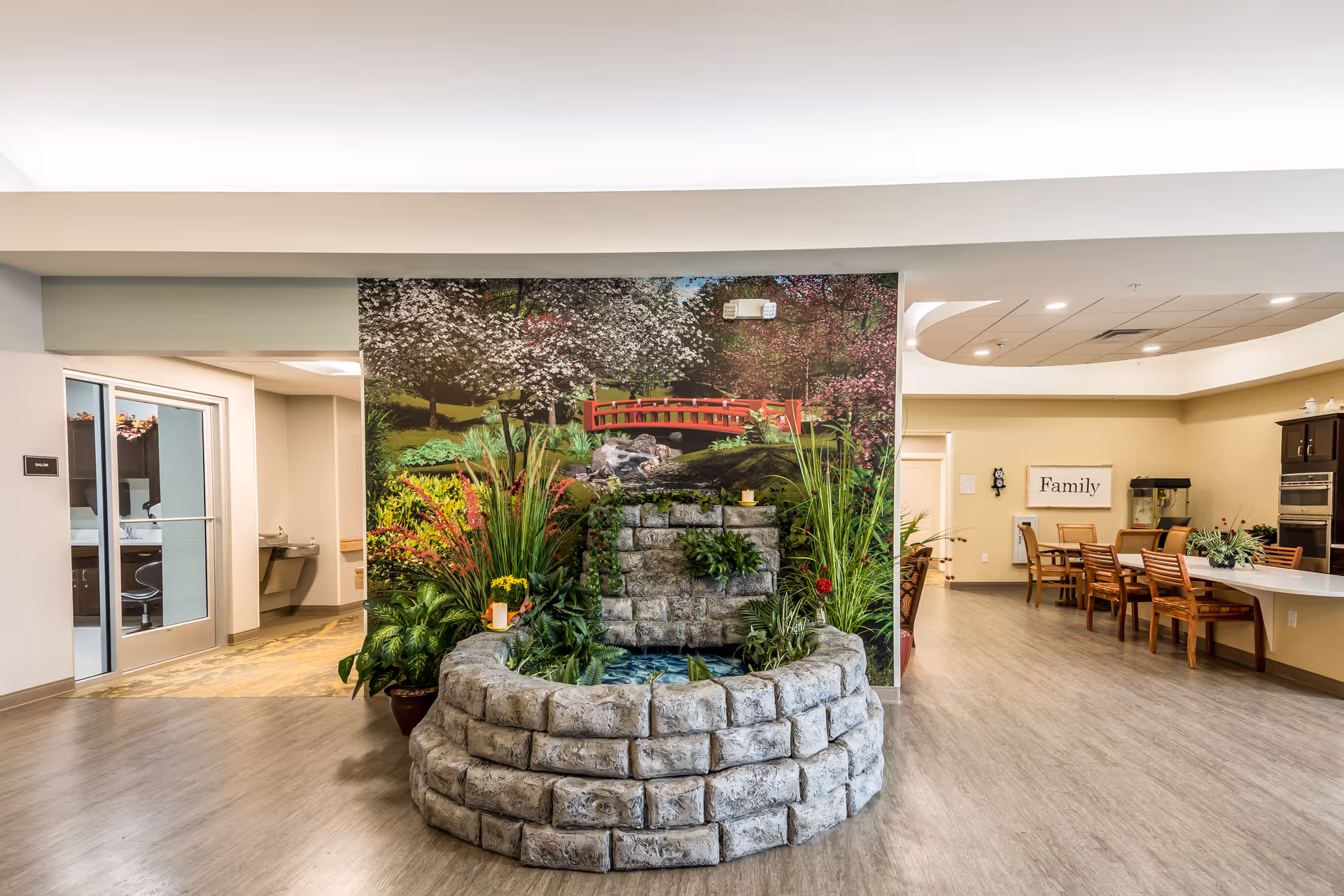 Interior view of a senior living facility common area featuring a decorative stone water fountain with plants in front of a mural depicting a garden with a red bridge. To the right, there is a dining area with wooden chairs and tables, and a wall sign that reads 'Family'. To the left, there is a hallway with a door leading to another room.