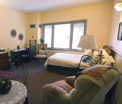 A cozy bedroom with a single bed near a window, a green armchair with a pillow, a wooden desk with a chair, decorative wall plates, and a table lamp on a nightstand.