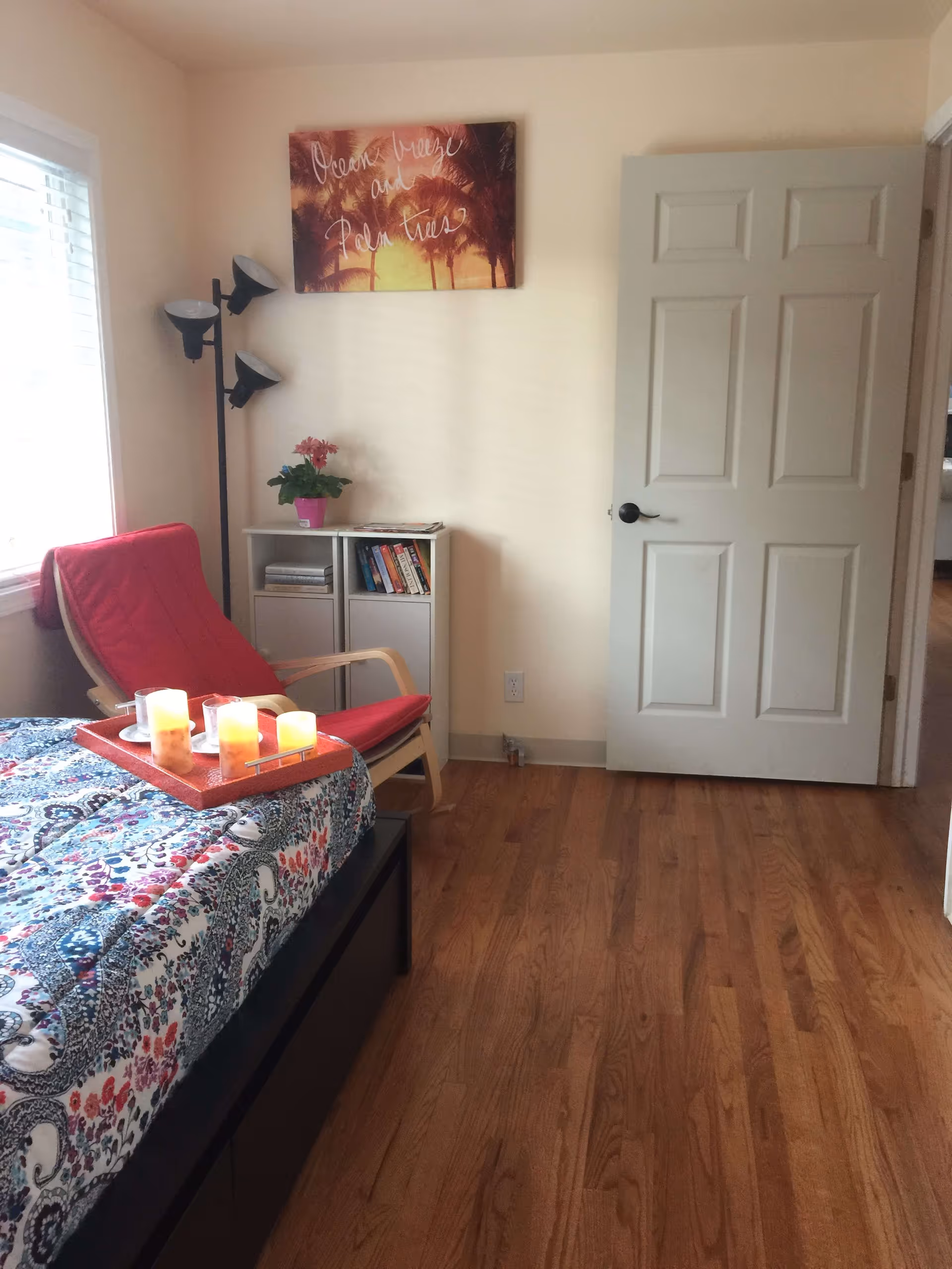 A bedroom with a bed covered in a colorful patterned quilt, a red cushioned chair next to a window with blinds, a small white bookshelf with books and a potted plant, a floor lamp with three adjustable lights, and a closed white door. A wall art above the bookshelf shows palm trees with the text 'Ocean breeze and Palm trees'.