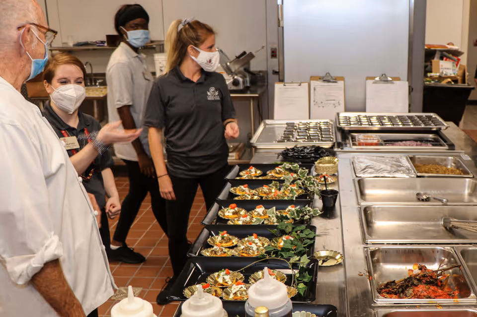 A group of four people wearing masks stand in a commercial kitchen near a counter with trays of prepared food. The kitchen has stainless steel surfaces and various food containers. The people appear to be discussing or observing the food preparation.