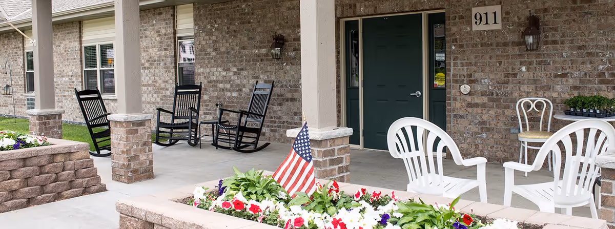 Front porch area of a brick building with a green door numbered 911. The porch has three black rocking chairs, two white plastic chairs, a small white table, and a flower bed with colorful flowers and an American flag in front.