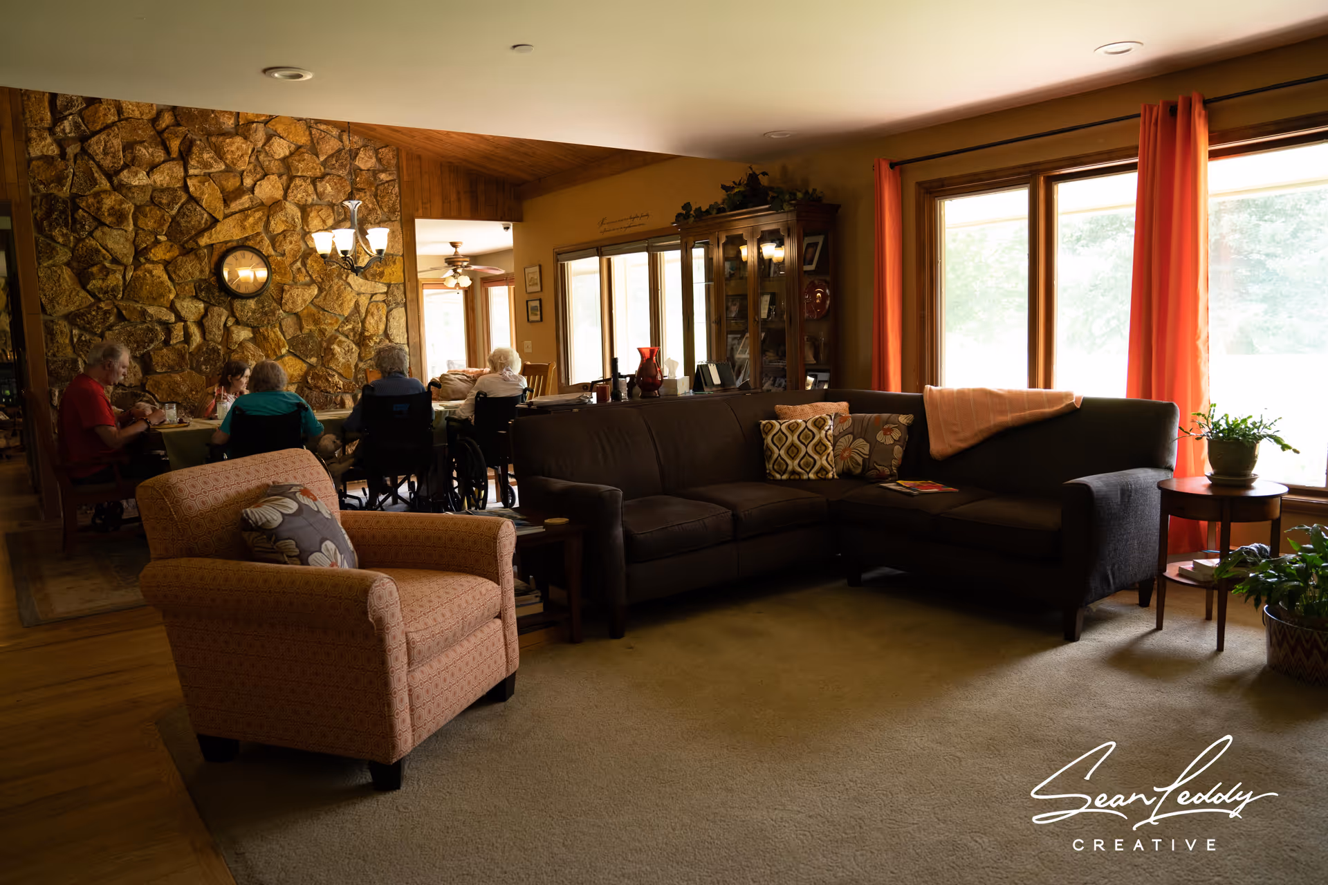 A cozy living room with a sectional sofa and armchair near large windows, with seniors seated at a dining table in the background.
