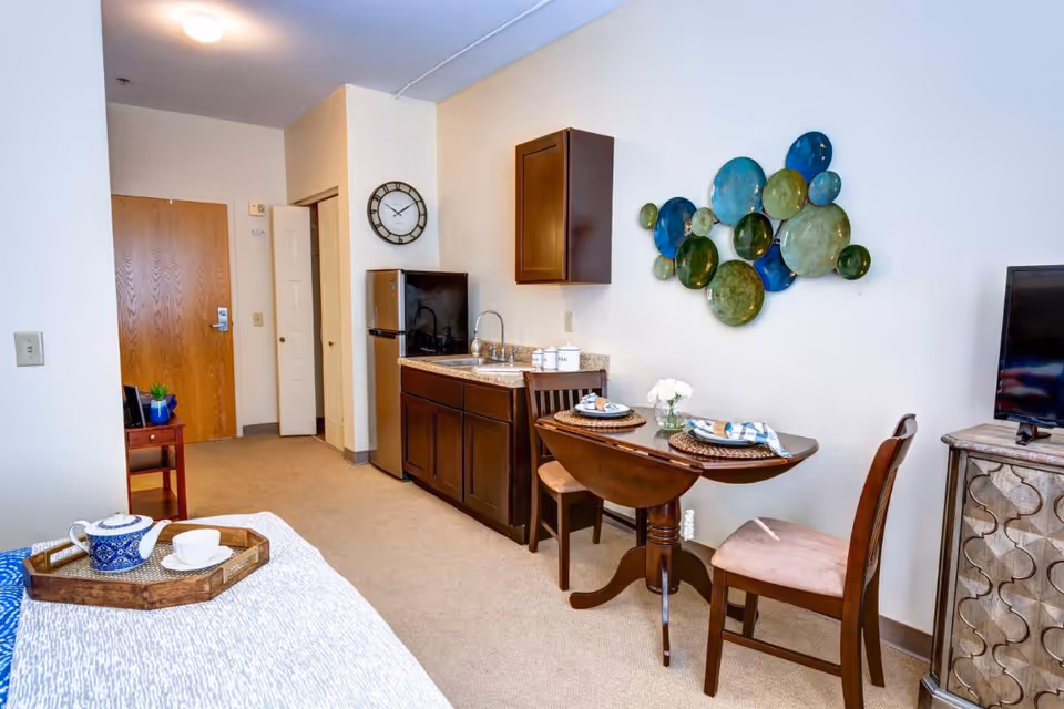 A cozy studio apartment interior featuring a small kitchenette with dark wood cabinets, a mini refrigerator, and a sink. A round wooden dining table with two chairs is set with placemats and napkins. Decorative wall art with blue and green circular glass pieces hangs above the table. A bed with a white and blue patterned blanket and a tray with a teapot and cup is partially visible. A TV sits on a wooden stand to the right, and a wooden door and closet are visible in the background.