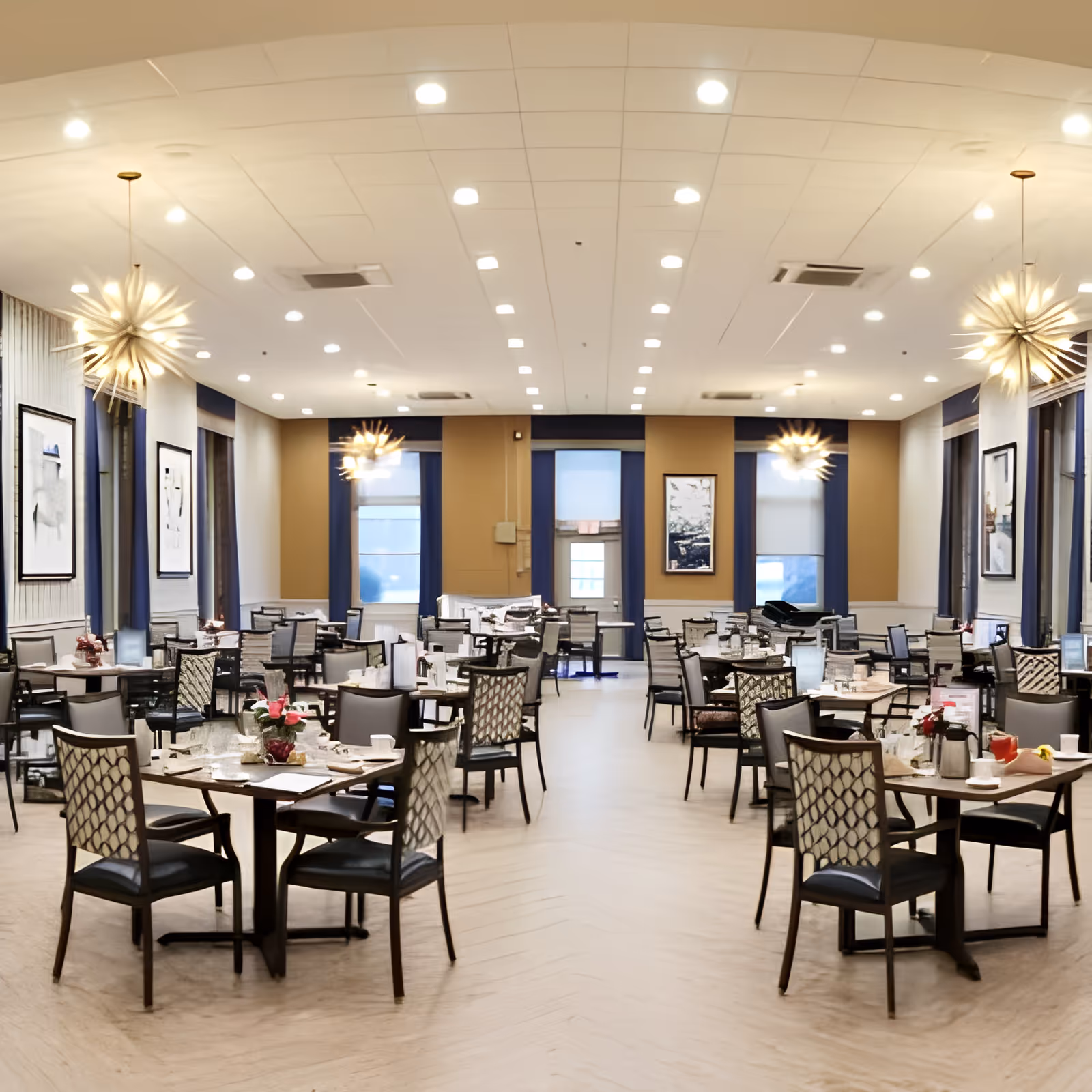 Spacious, well-lit dining room with multiple set tables, patterned chairs, and modern starburst chandeliers.