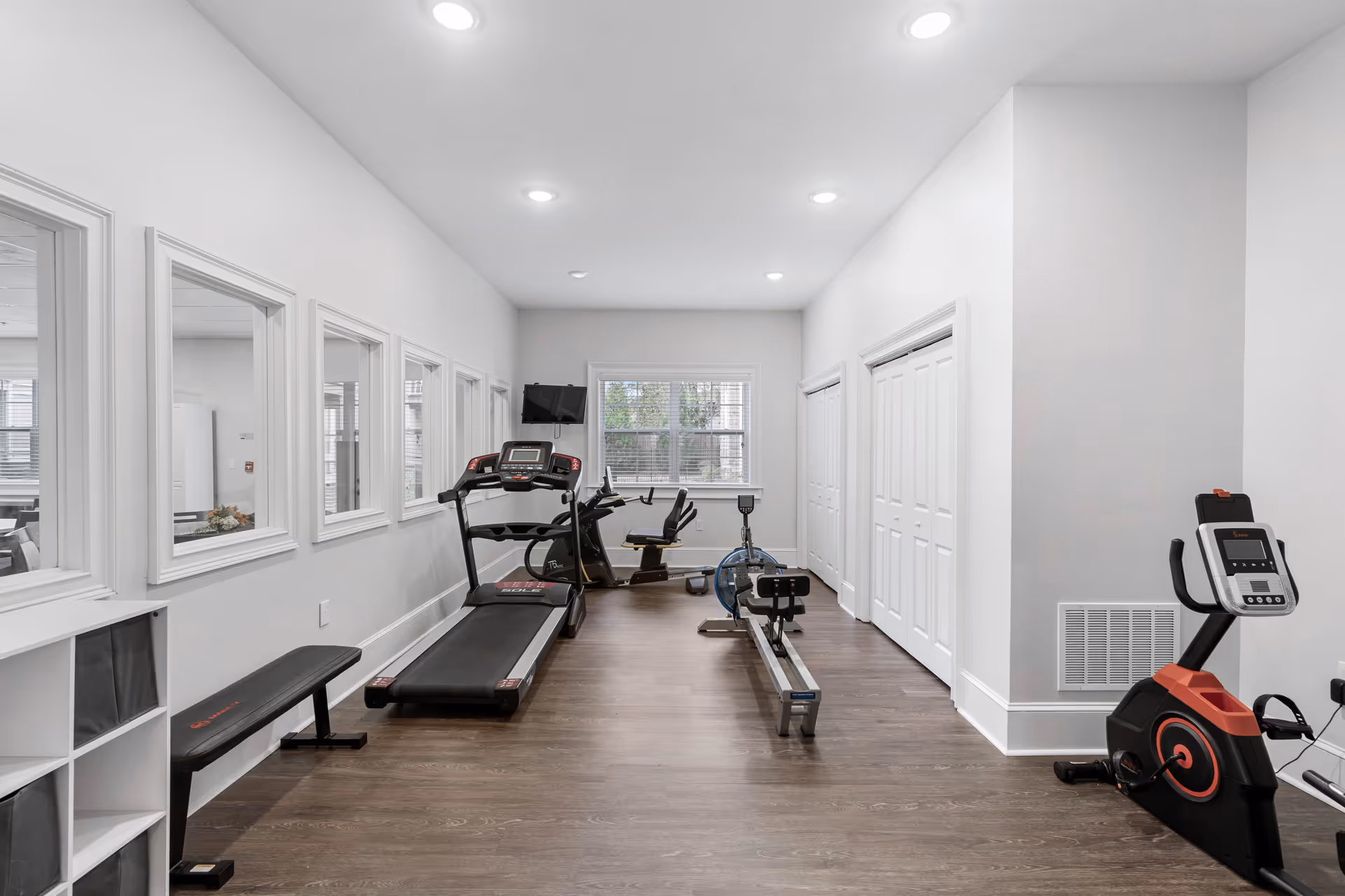 A small fitness room with exercise equipment including a treadmill, rowing machine, stationary bike, and a bench. The room has white walls, wooden flooring, recessed ceiling lights, and a window at the far end letting in natural light. Several windows on the left wall look into an adjacent room.