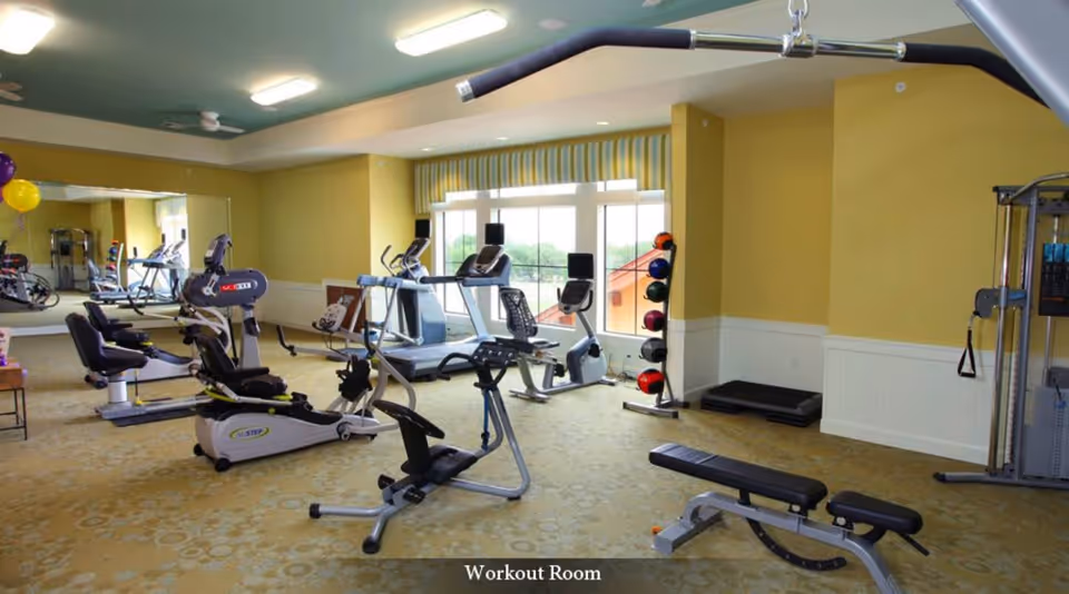 A workout room in a senior living facility with various exercise equipment including stationary bikes, treadmills, a weight bench, and a rack of medicine balls. The room has yellow walls, a large window with striped valance, and a large mirror on one wall.