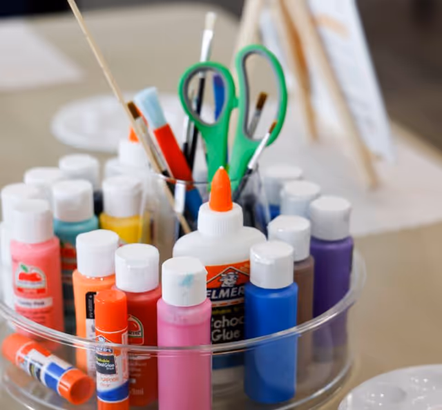 Clear organizer holding bottles of colorful craft paint, glue, glue sticks, brushes, and scissors on a table.