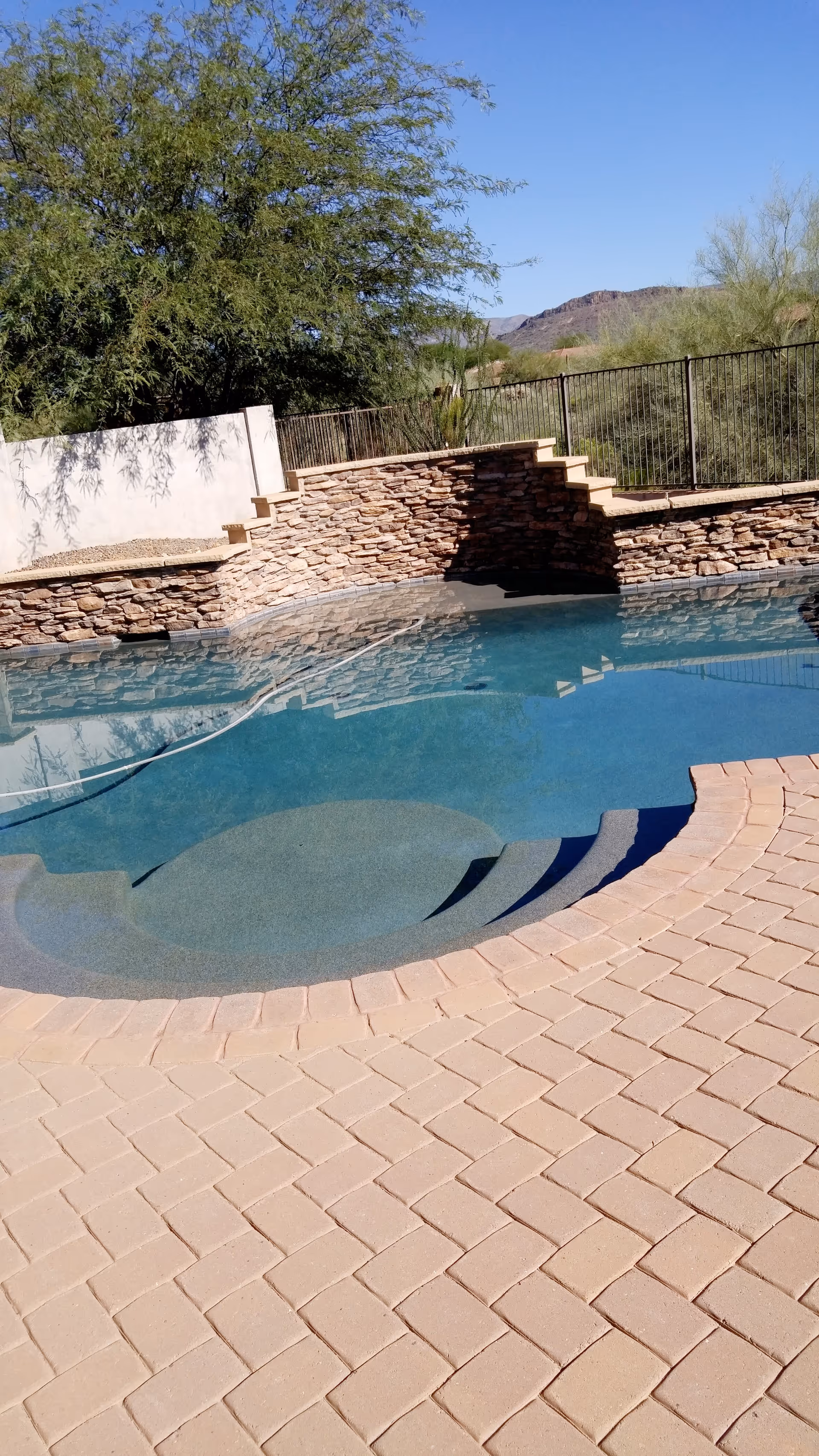 Outdoor swimming pool with curved steps leading into the water, surrounded by a stone wall and a paved patio area. There are trees and desert vegetation in the background under a clear blue sky.