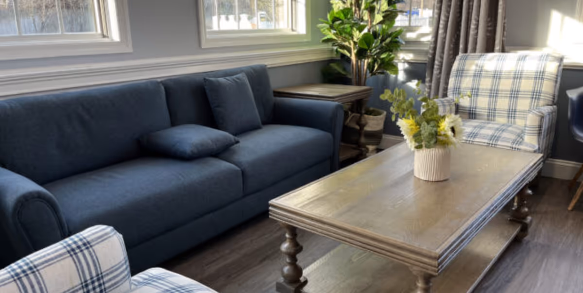 Bright living room with a blue sofa, plaid armchairs, a wooden coffee table with a vase of flowers, and windows letting in daylight.