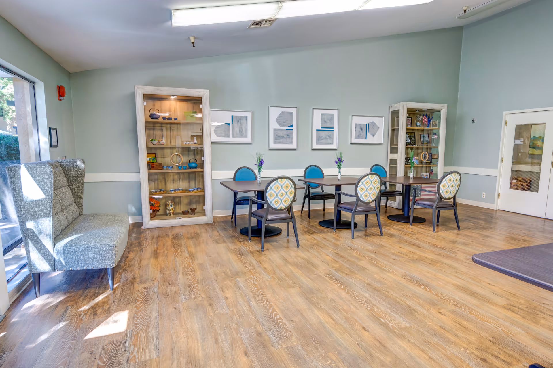 Bright assisted living common room with round tables and patterned chairs, display cabinets, and a bench by the window.