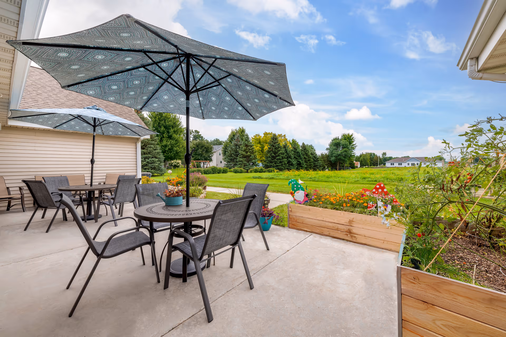 Outdoor patio area with round metal tables and chairs under large patterned umbrellas. Raised wooden garden beds with flowers and decorative garden ornaments are visible, along with a grassy field and trees in the background under a partly cloudy sky.