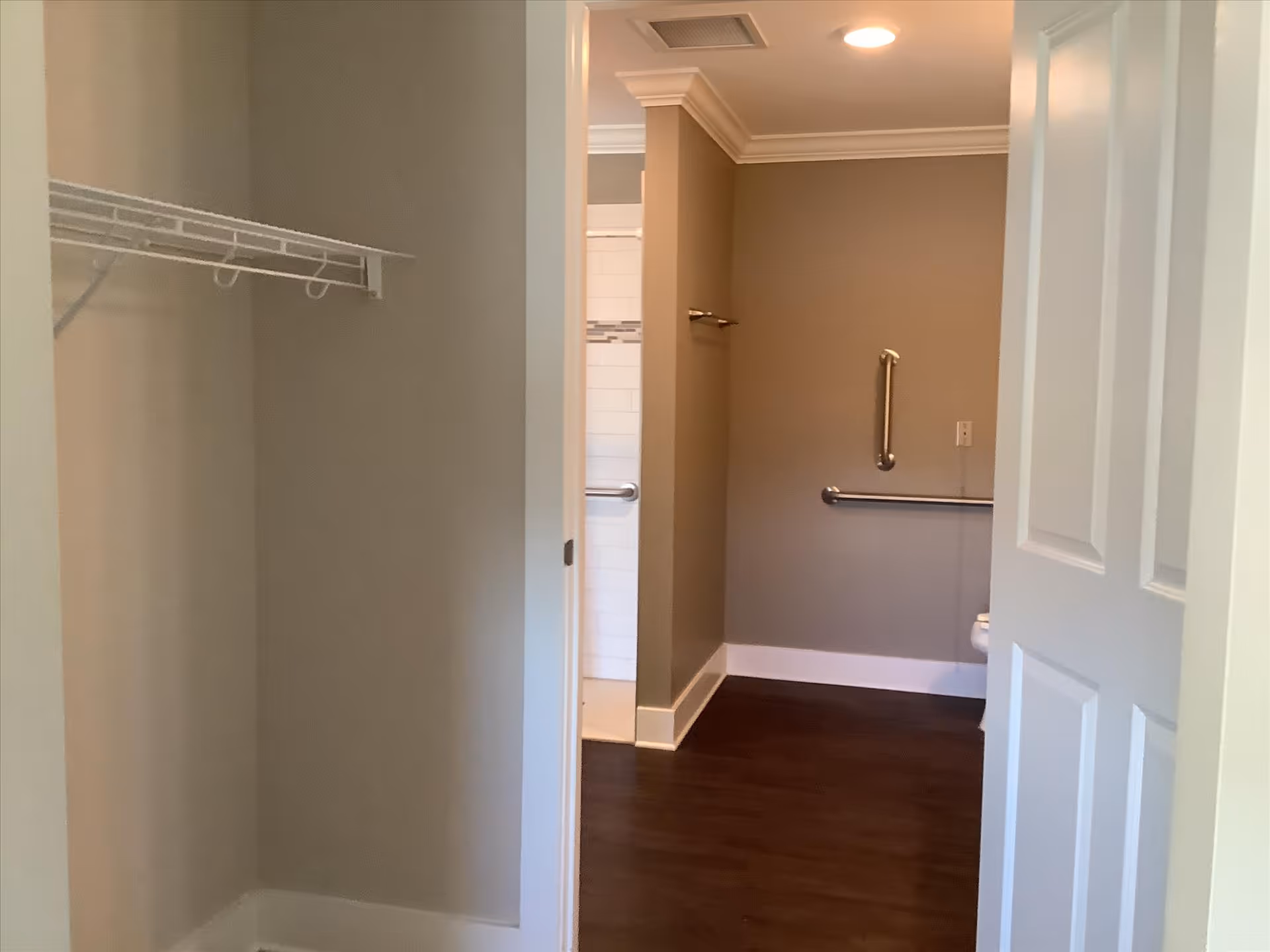 Interior view from a closet into an accessible bathroom with grab bars, a tiled shower entrance, and a partially open door.