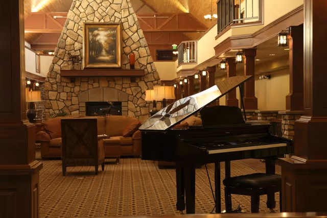 A cozy living room area with a grand piano in the foreground, a stone fireplace with a framed painting above it, and comfortable seating including sofas and chairs. The room features warm lighting, wooden beams, and a patterned carpet.