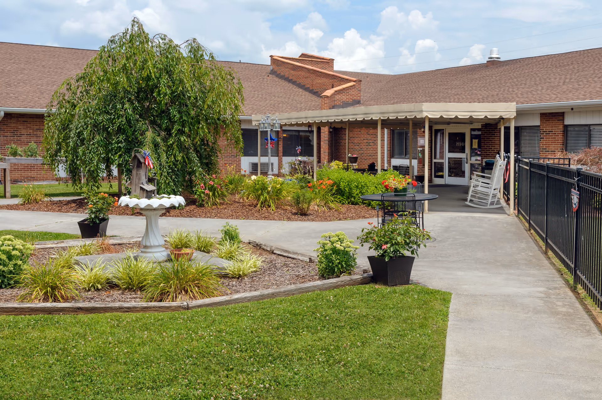 Outdoor view of a senior living facility with a brick building, a covered entrance with rocking chairs, a paved walkway, and a landscaped garden area featuring a birdbath and various plants under a partly cloudy sky.